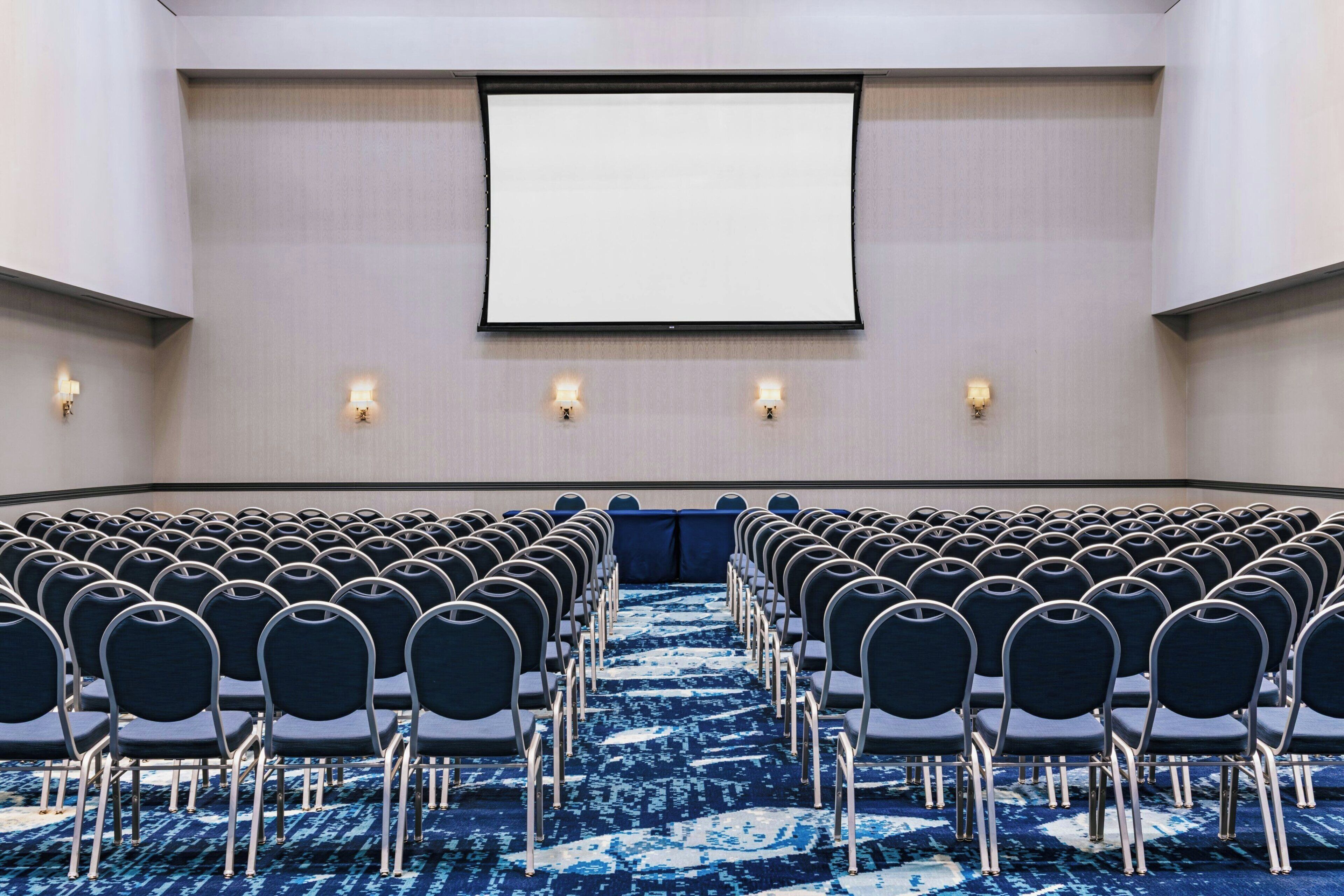 A conference room with rows of chairs, a large screen, and wall-mounted lights.