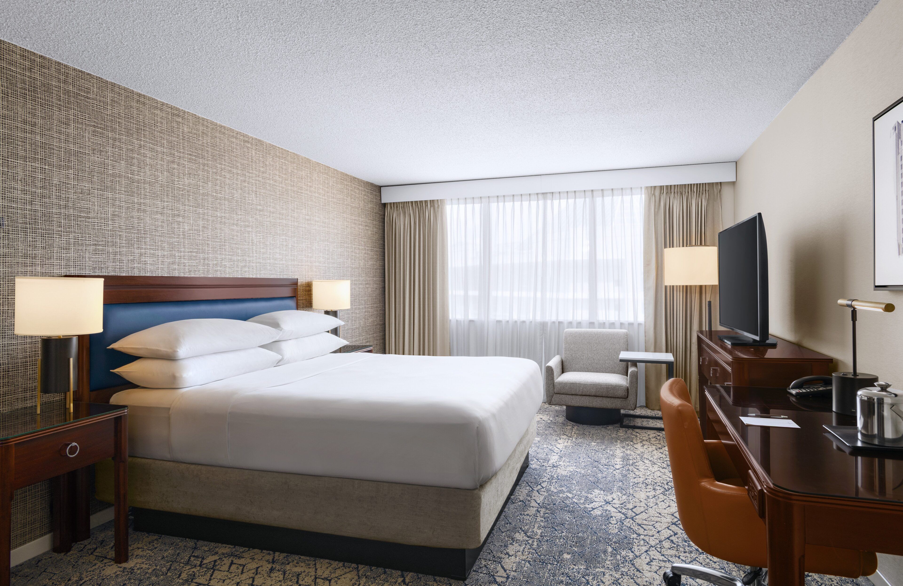 A hotel room with a large bed, a desk, a chair, a television, and a window with curtains.