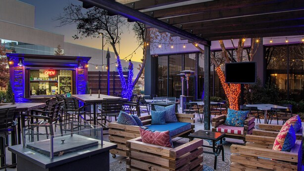 Un área de estar al aire libre con mobiliario de madera y un cerramiento de vidrio, un restaurante con el letrero 'OPEN' y luces colgantes.