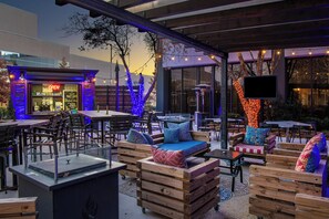Un área de estar al aire libre con mobiliario de madera y un cerramiento de vidrio, un restaurante con el letrero 'OPEN' y luces colgantes.