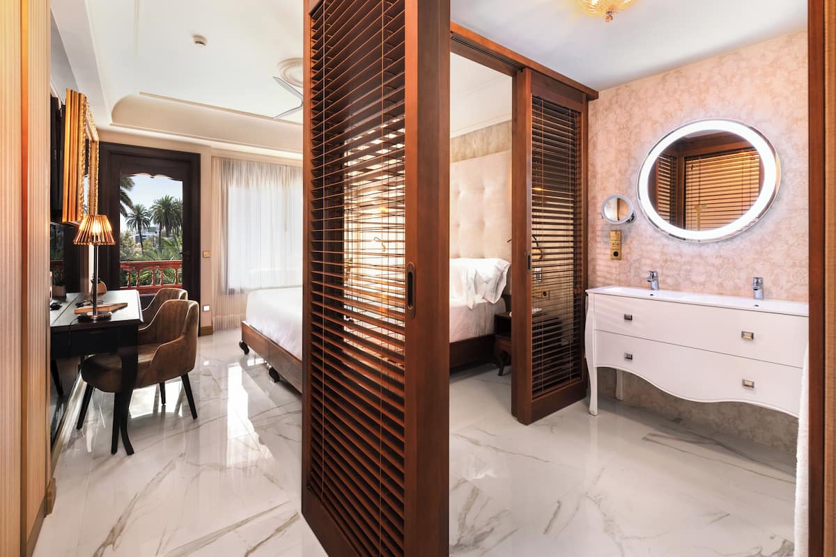 Habitación de hotel con cama, escritorio con sillas, baño con tocador y espejo, y vista al exterior.