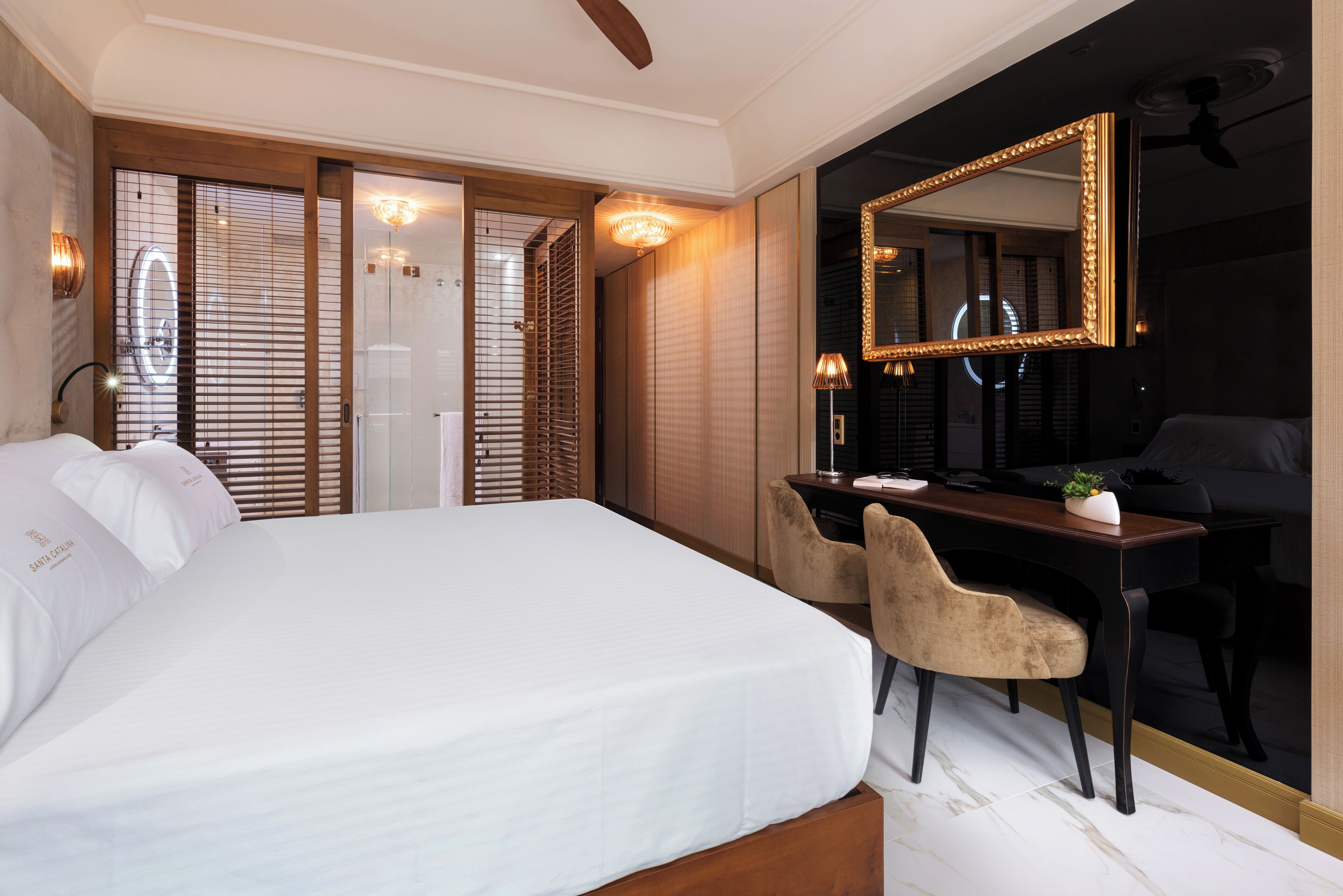 A hotel room with a large bed, a desk with a chair, a mirror, and a shower area.