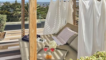 A balcony with a wooden pergola, white curtains, a cushioned seating area, and a tray with drinks and snacks.