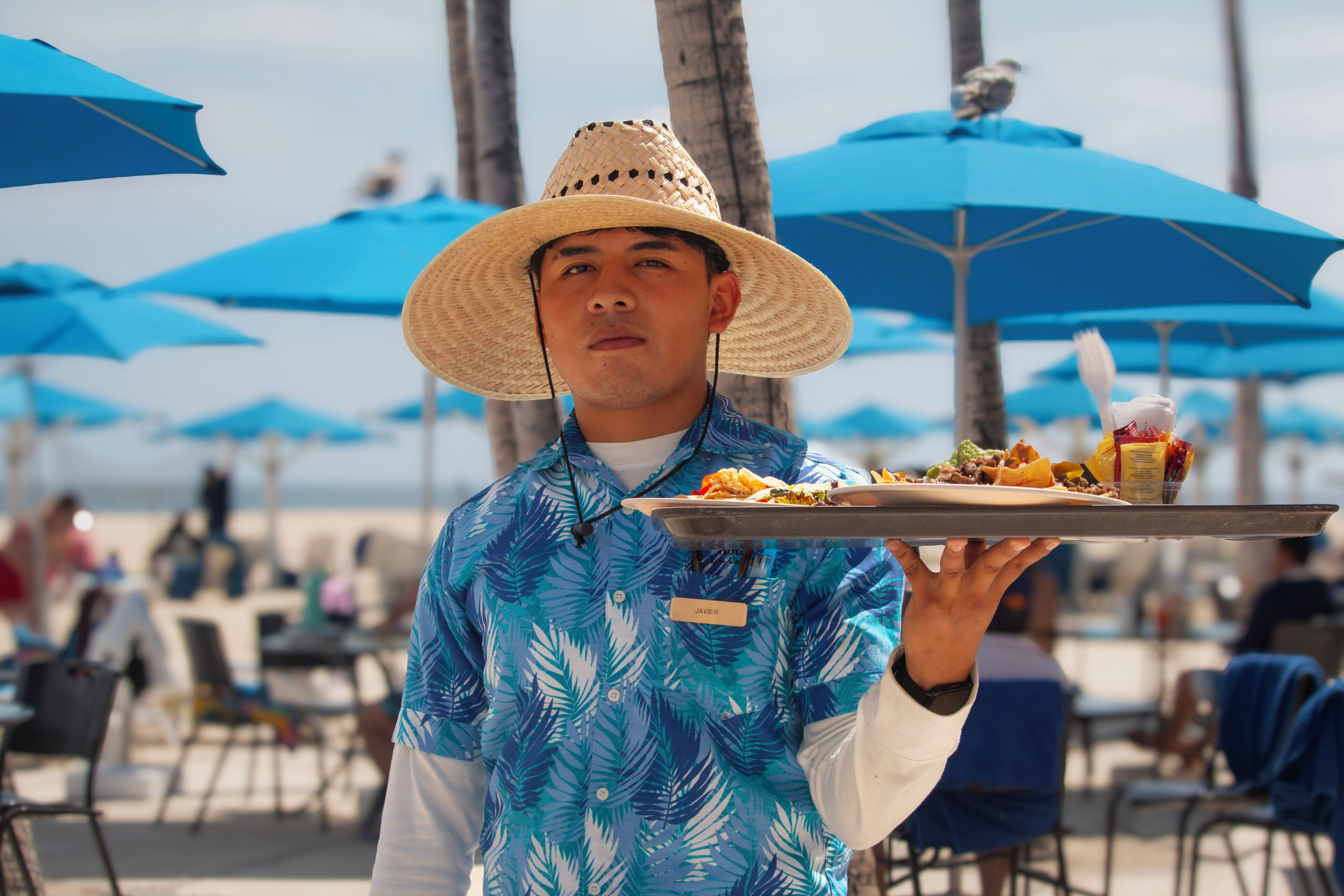 A person wearing a straw hat and a blue shirt with palm leaf patterns, holding a tray with food and condiments.