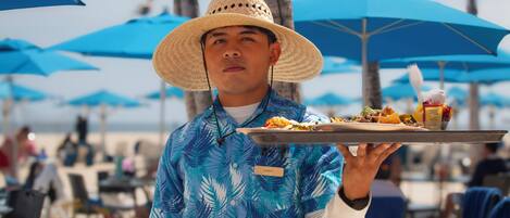 A person wearing a straw hat and a blue shirt with palm leaf patterns, holding a tray with food and condiments.