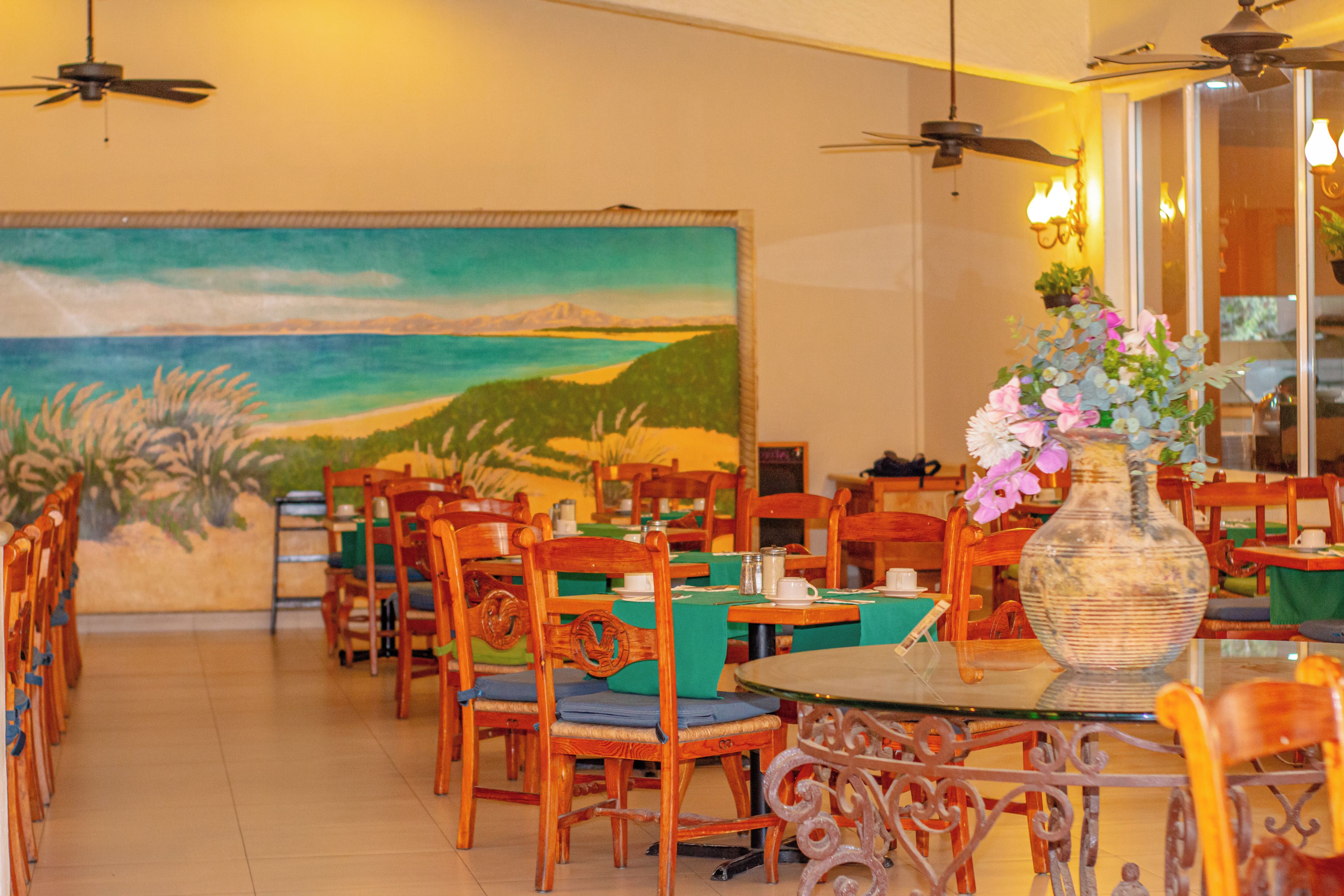 Interior de restaurante con sillas y mesas de madera, y un gran mural con una escena de playa en la pared.