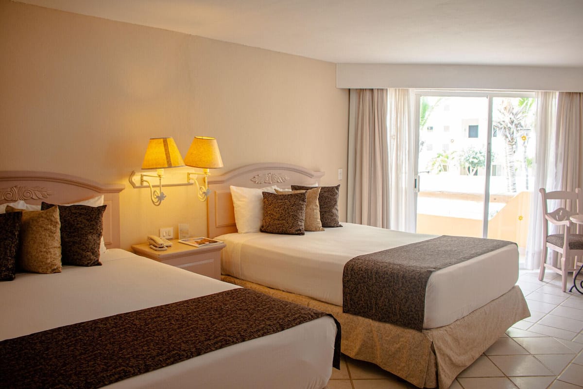A hotel room with two beds, a nightstand with a lamp, a chair, and a view of the outside.