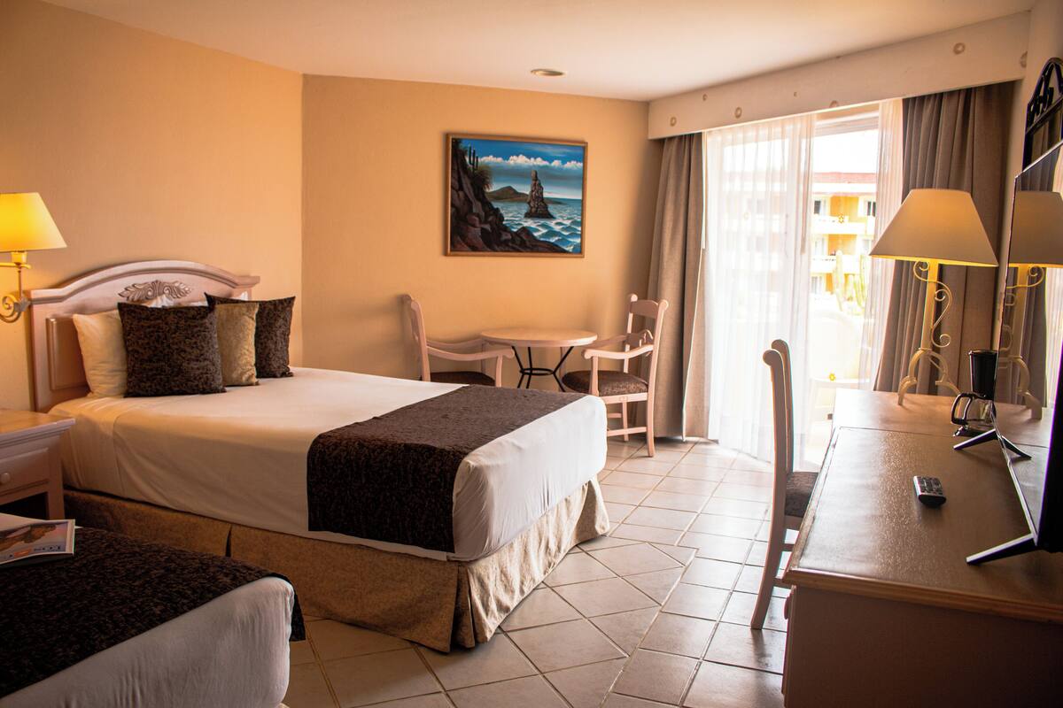 A hotel room with a bed, a desk, a chair, a lamp, and a painting on the wall.