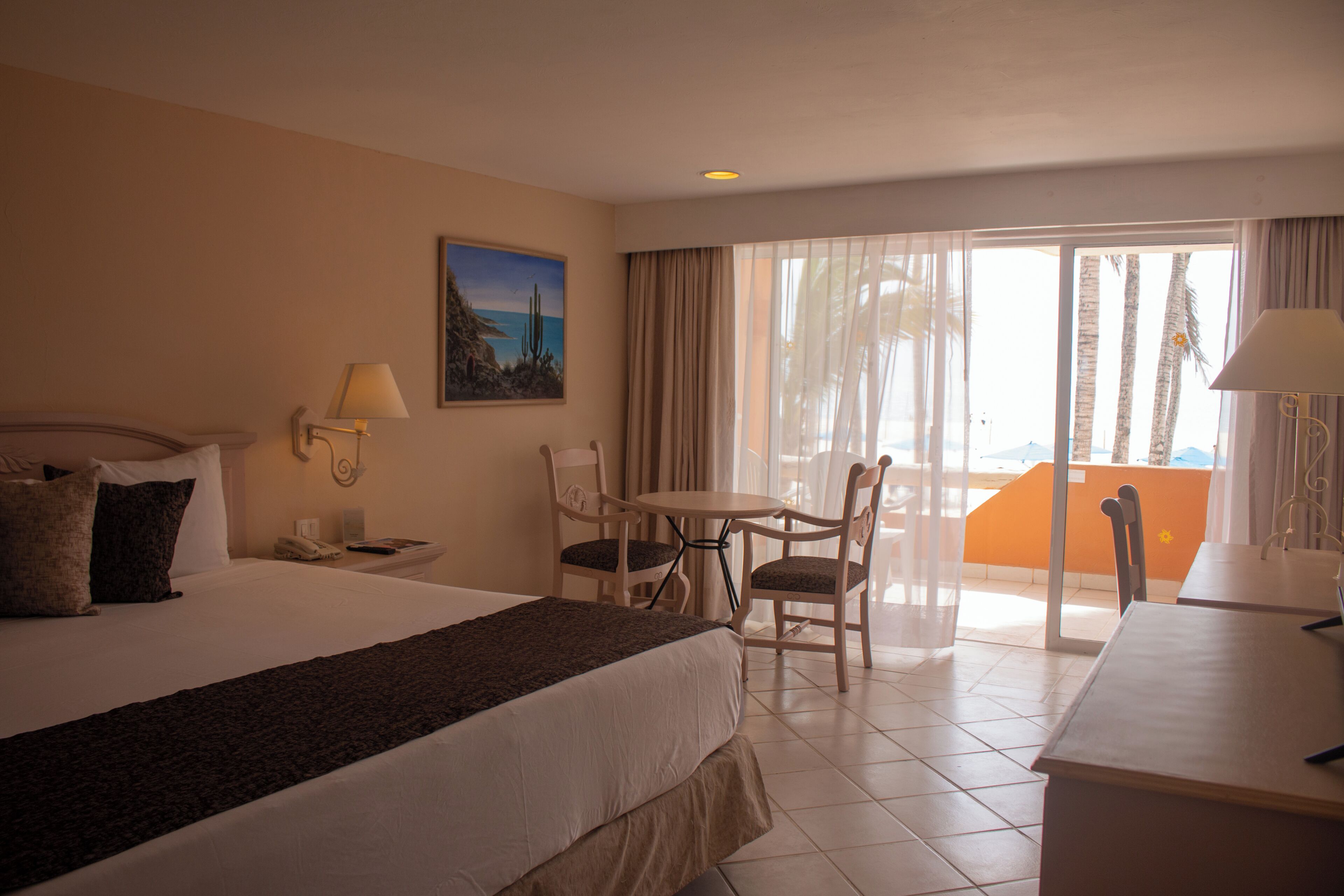A hotel room with a bed, a desk, a chair, a table, a painting, and a balcony with a view.