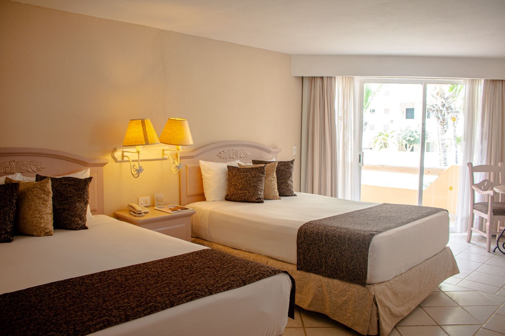 A hotel room with two beds, a nightstand with a lamp, a chair, and a view of the outdoors.