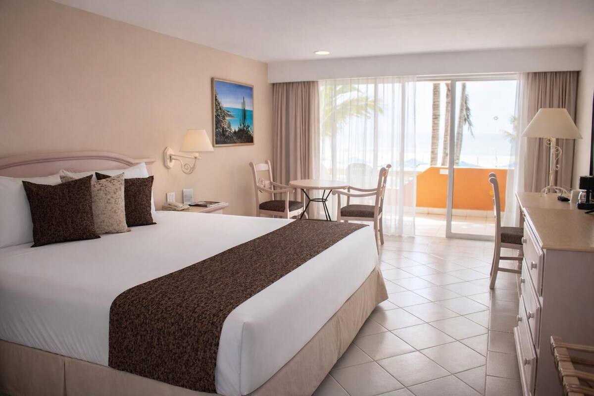 A hotel room with a large bed, a desk, a chair, a balcony with curtains, and a painting on the wall.