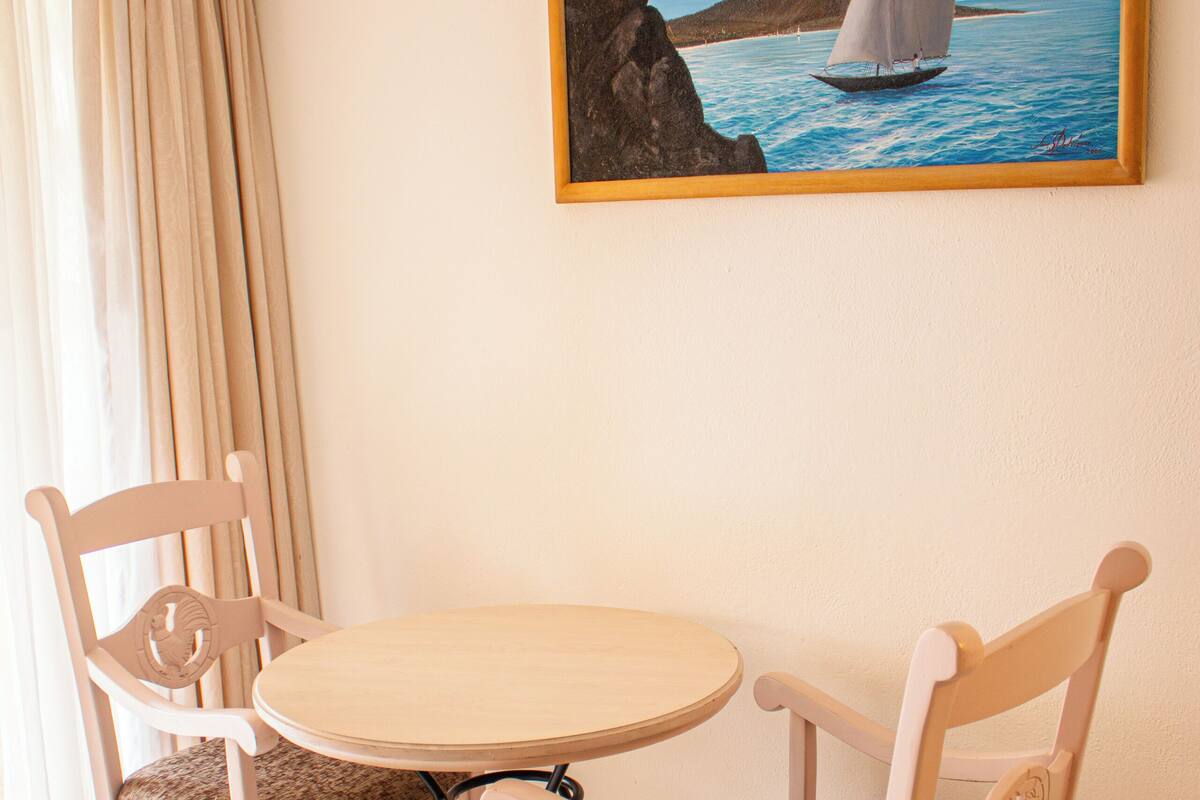 A dining area with a table and chairs, a painting of a sailboat on the wall, and a window with curtains.