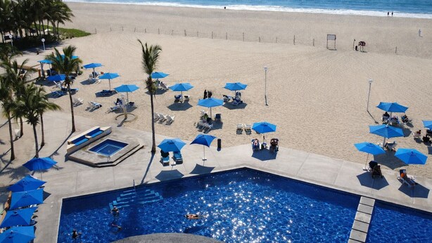 A pool area with blue umbrellas, lounge chairs, and a beach in the background.