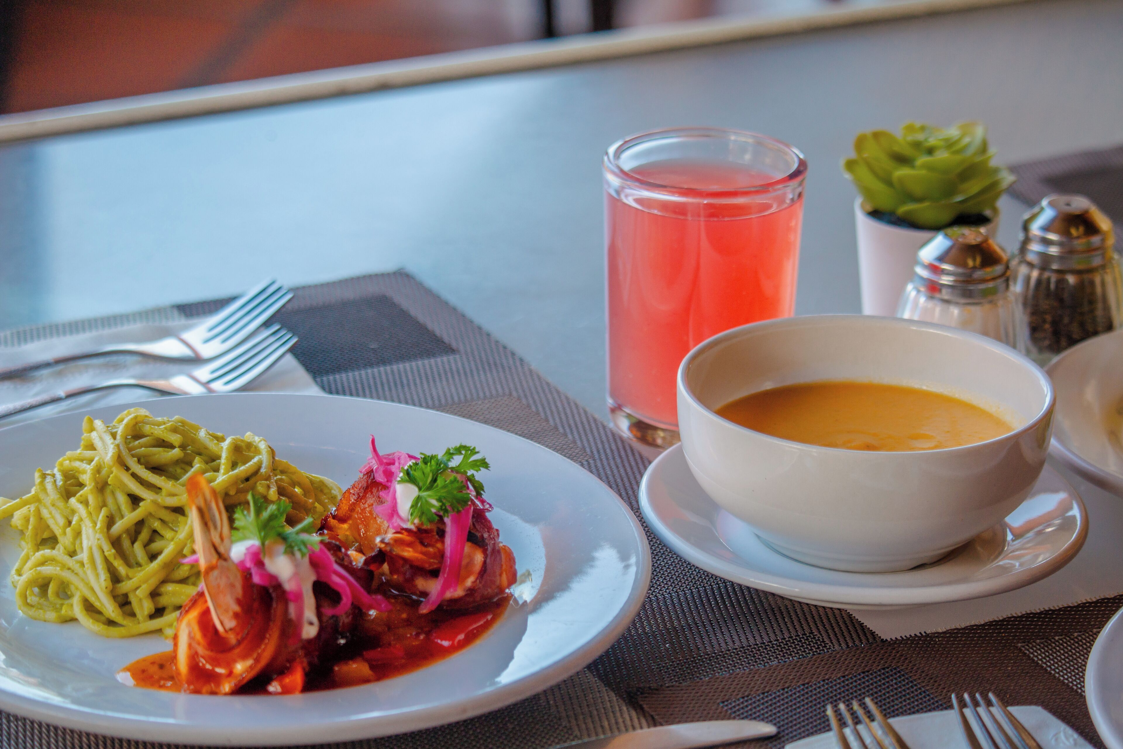 Une table à manger avec une assiette de pâtes, un bol de soupe, un verre d’une boisson rose et une petite plante.