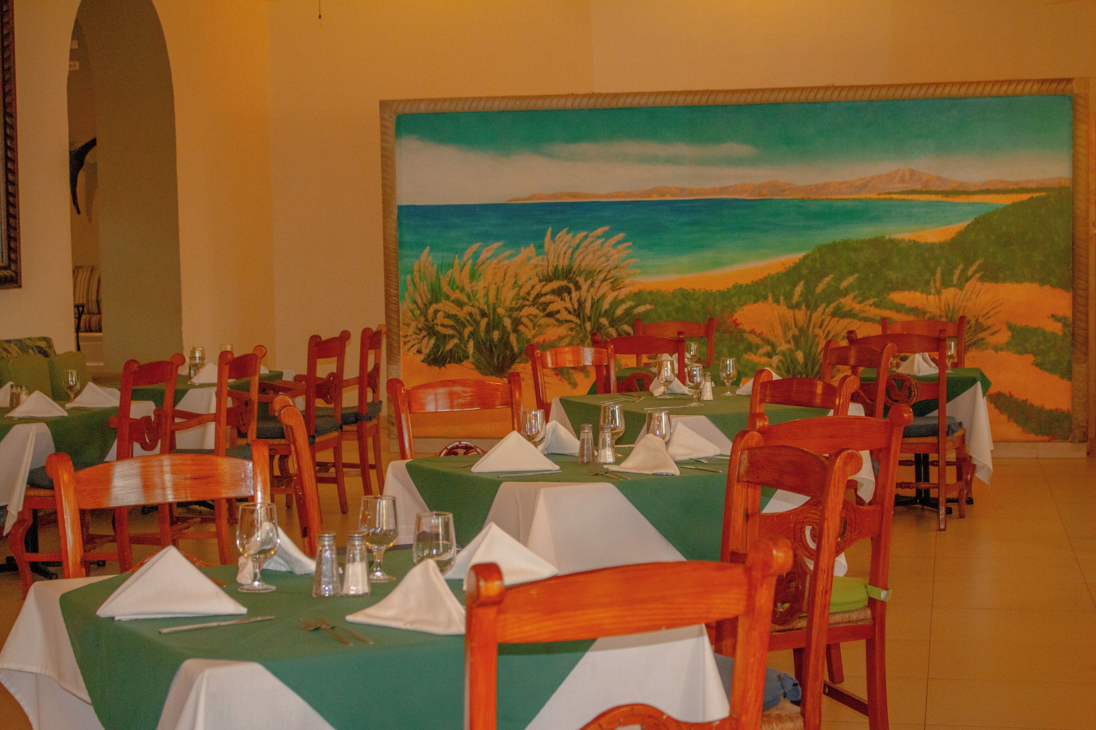 Un comedor con mesas dispuestas para el servicio, sillas y un cuadro grande de un paisaje costero.