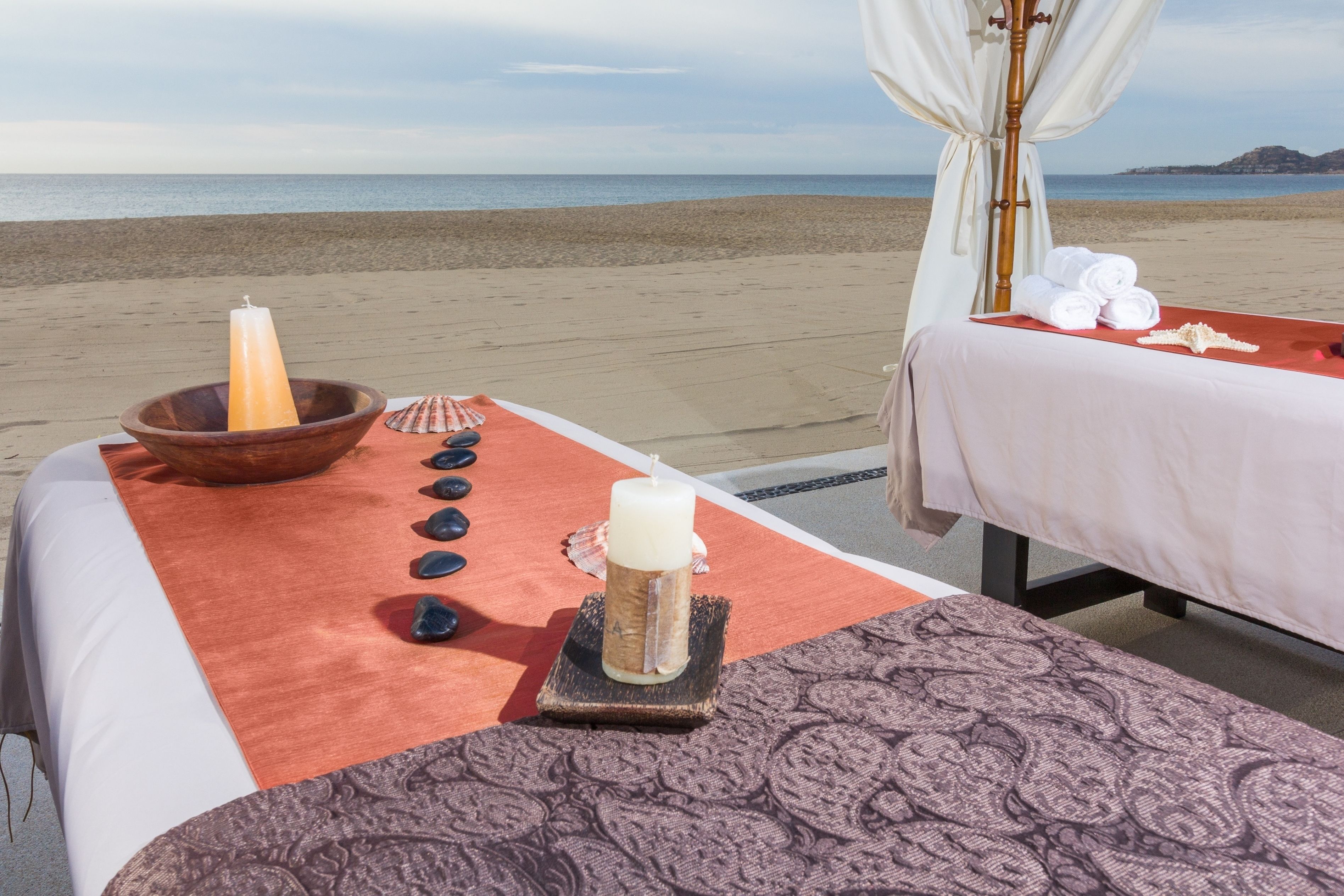 Un espace de bien-être aménagé avec une table de massage, une bougie et un récipient, le tout installé sur une plage avec vue sur la mer.