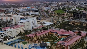 A coastal resort with multiple hotel buildings, a sandy beach with umbrellas, and a view of the mountains in the background.