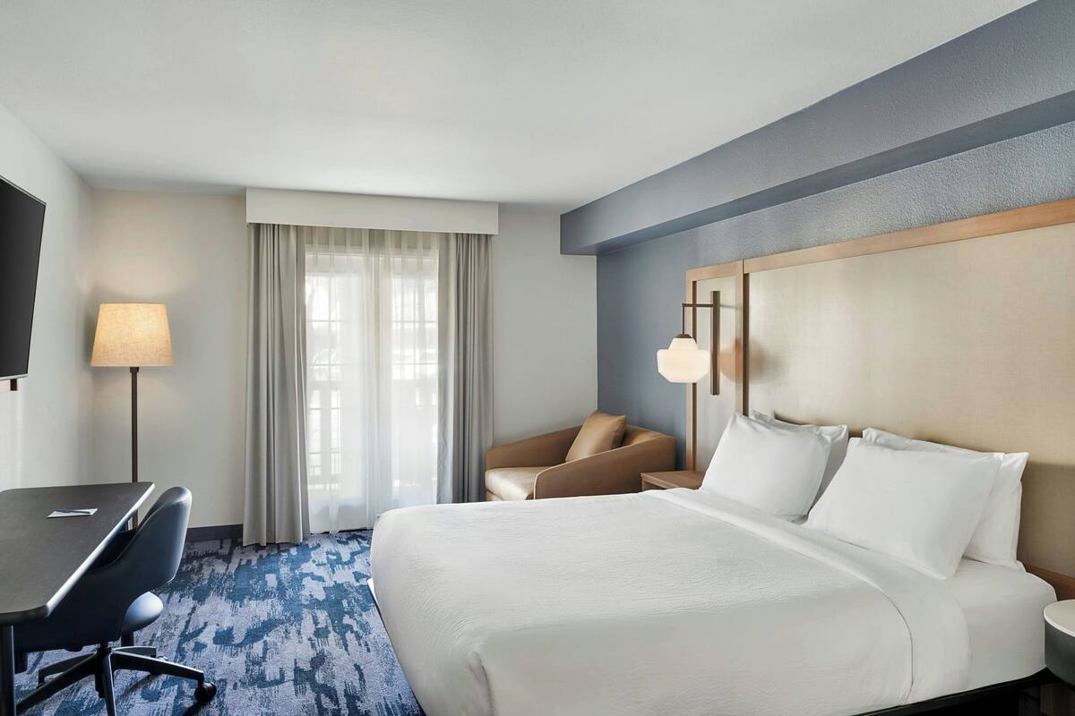 Ein Hotelzimmer mit Bett, Schreibtisch, Stuhl, Fernseher und einem fensterbepflanzten Fenster.