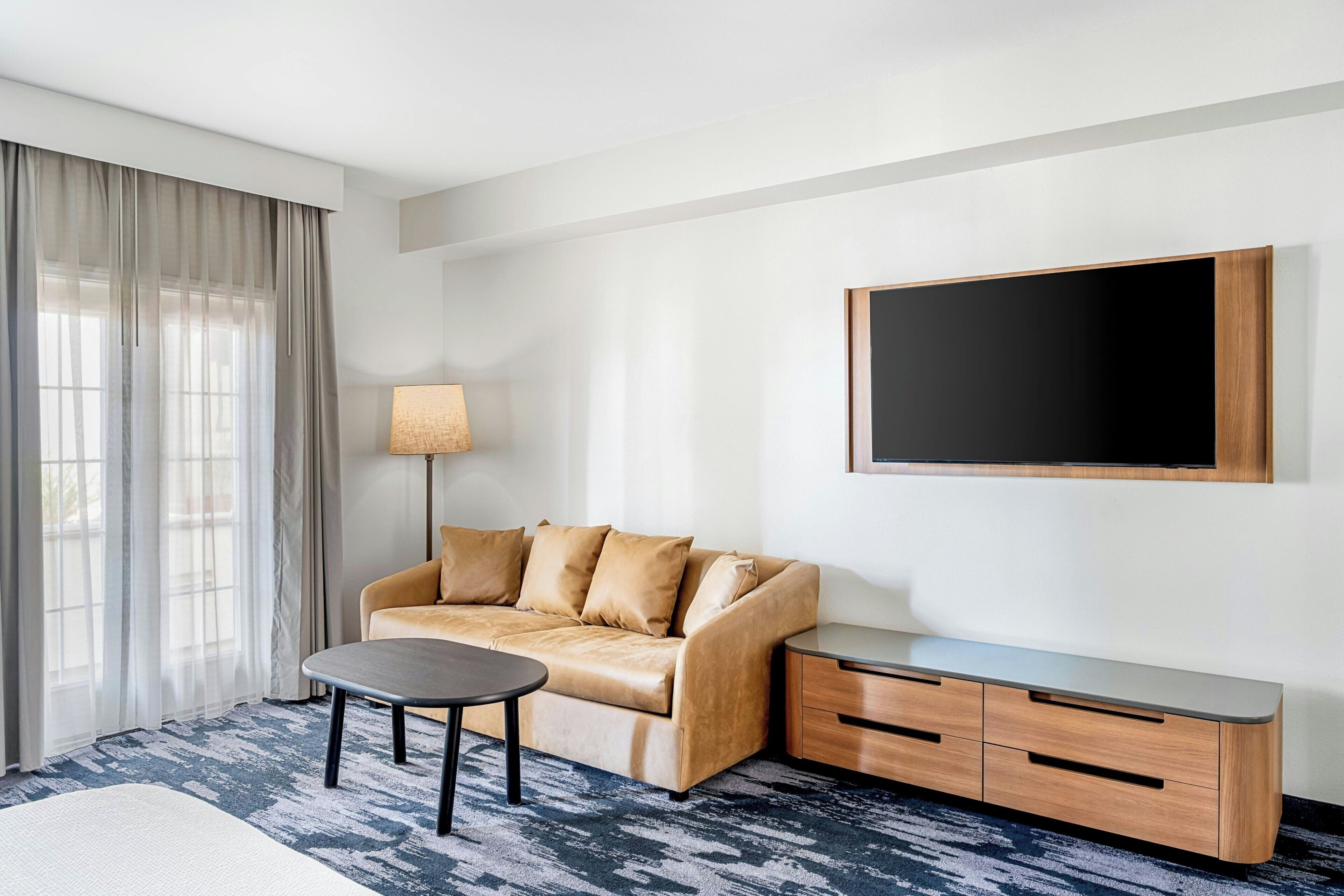 Quarto de hotel com sofá, mesa pequena, televisão na parede e janela com cortinas.