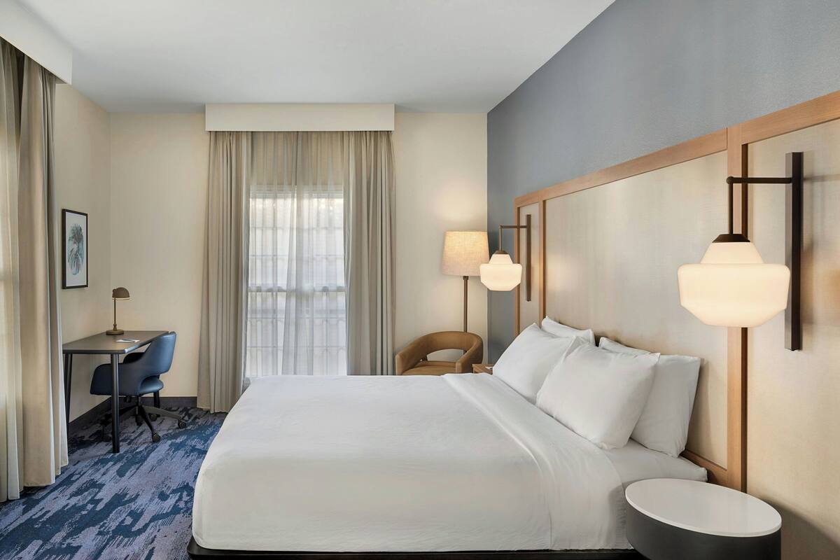 Ein Hotelzimmer mit einem großen Bett, einem Schreibtisch mit Stuhl, einer Lampe und einem Fenster mit Vorhängen.