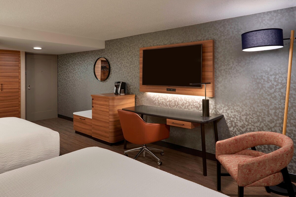 Quarto de hotel com cama, escrivaninha, cadeira, televisão e abajur.