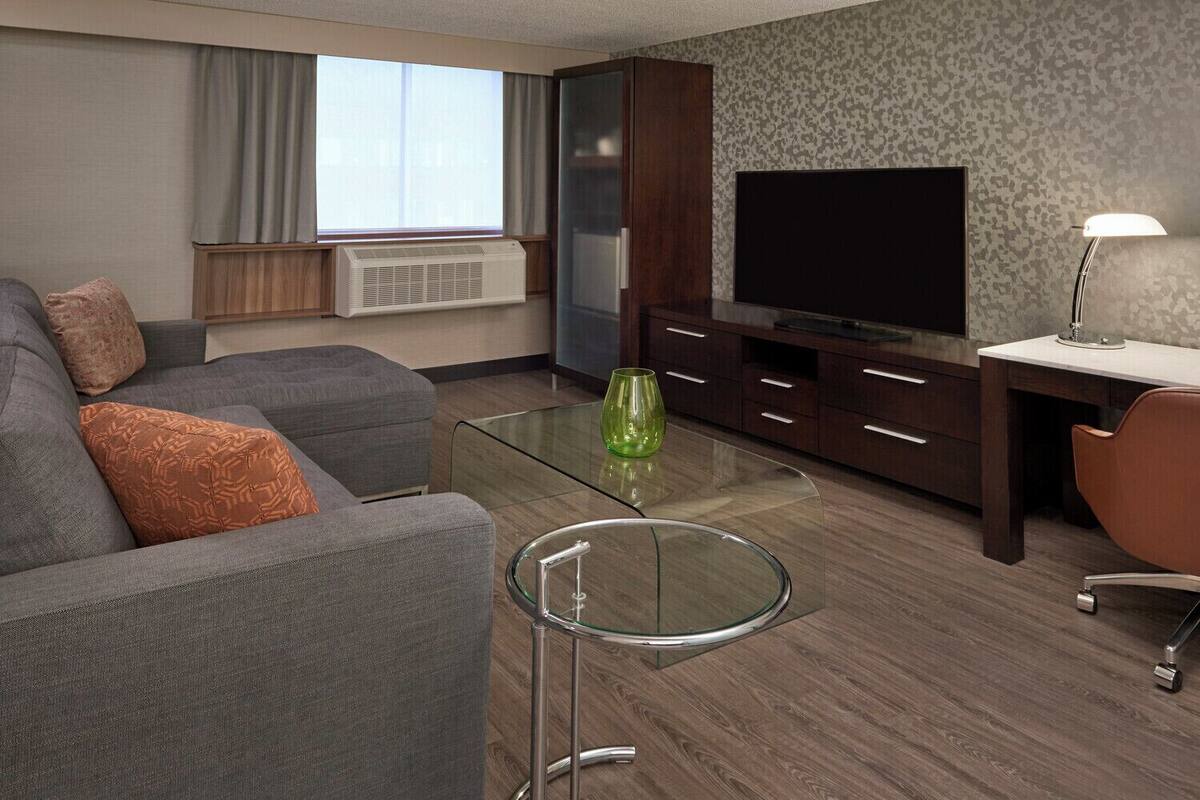 Et moderne hotelværelse med en grå sofa, et glasbord, et fjernsyn og et skrivebord med en lampe.