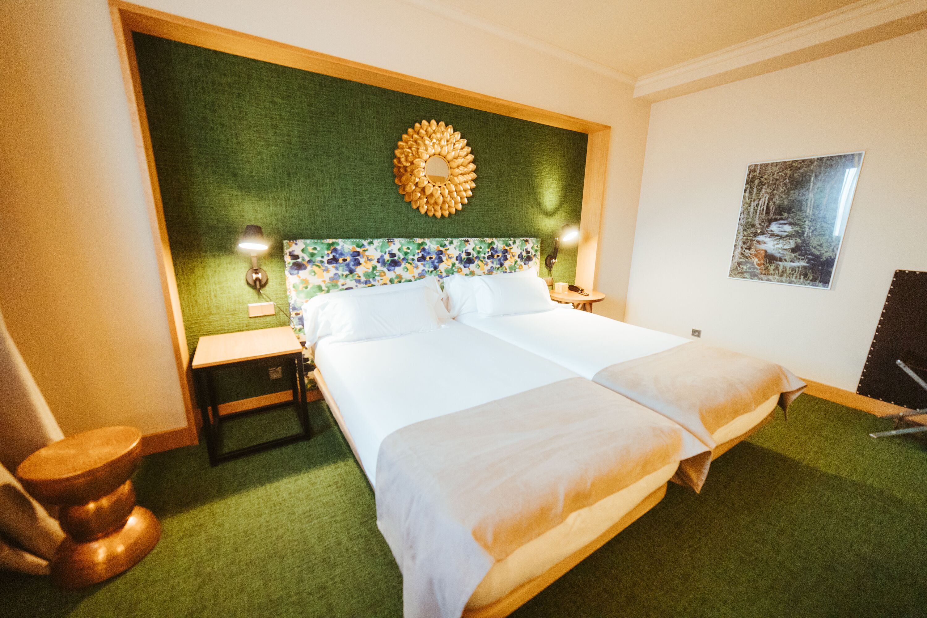 A hotel room with a large bed, a green headboard, a decorative mirror, a nightstand, a lamp, and a framed picture on the wall.