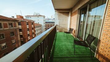 A balcony with a green artificial turf, two black chairs, and a view of surrounding buildings.
