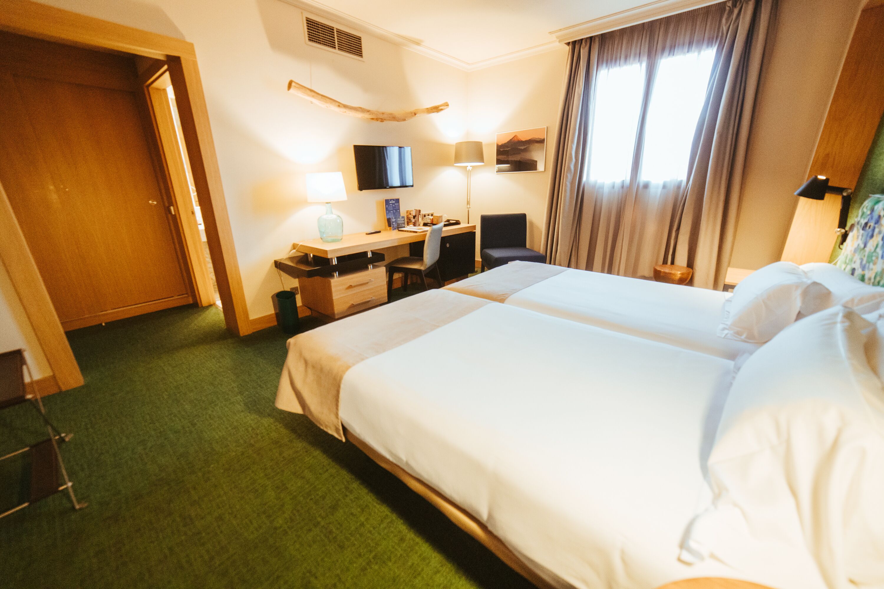 A hotel room with a large bed, a desk with a chair, a television, and a window with curtains.