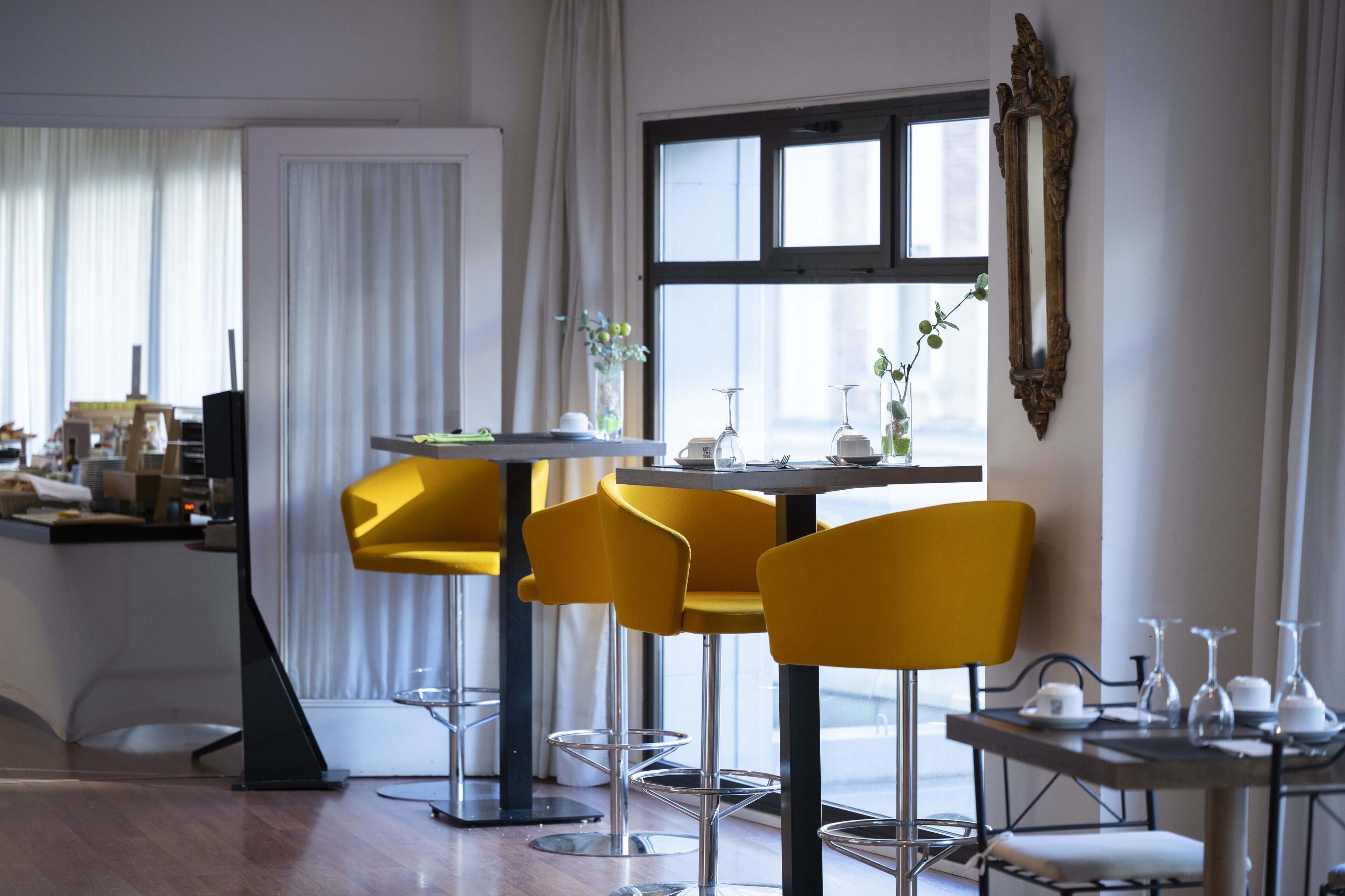 A modern dining area with a table set for four, surrounded by yellow chairs, a decorative mirror, and a window with curtains.