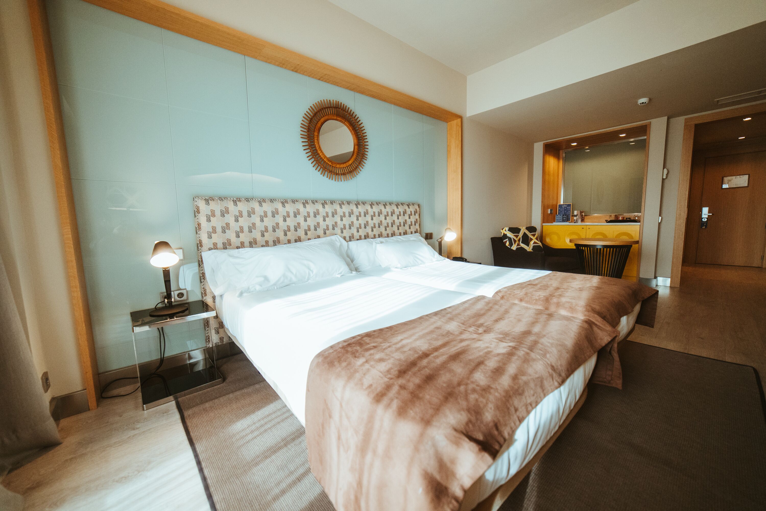 A hotel room with a large bed, a bedside table with a lamp, a mirror above the bed, and a small kitchenette area.