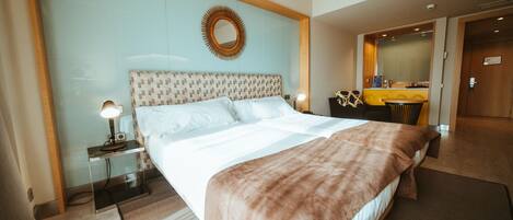 A hotel room with a large bed, a bedside table with a lamp, a mirror above the bed, and a small kitchenette area.