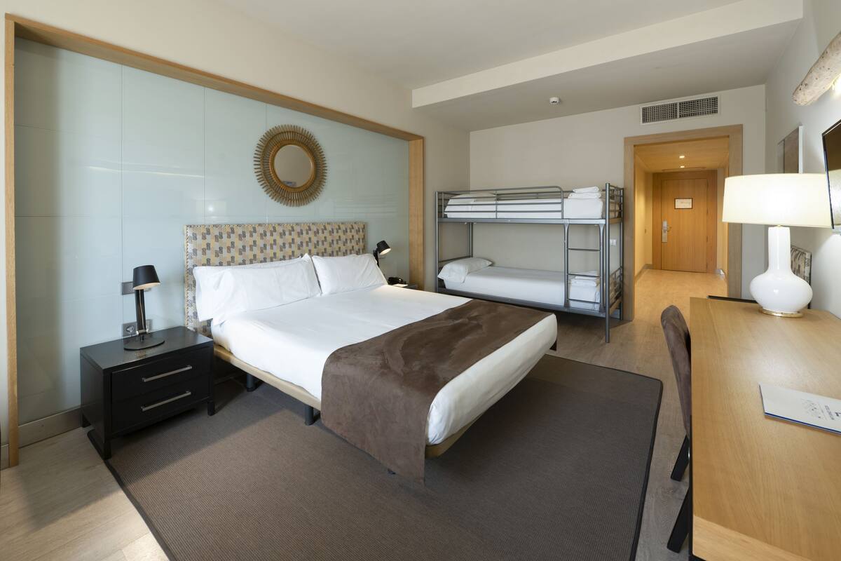 Habitación de hotel con una cama grande, una mesita de noche con lámpara, una cama nido y un escritorio con lámpara, además de un espejo en la pared.