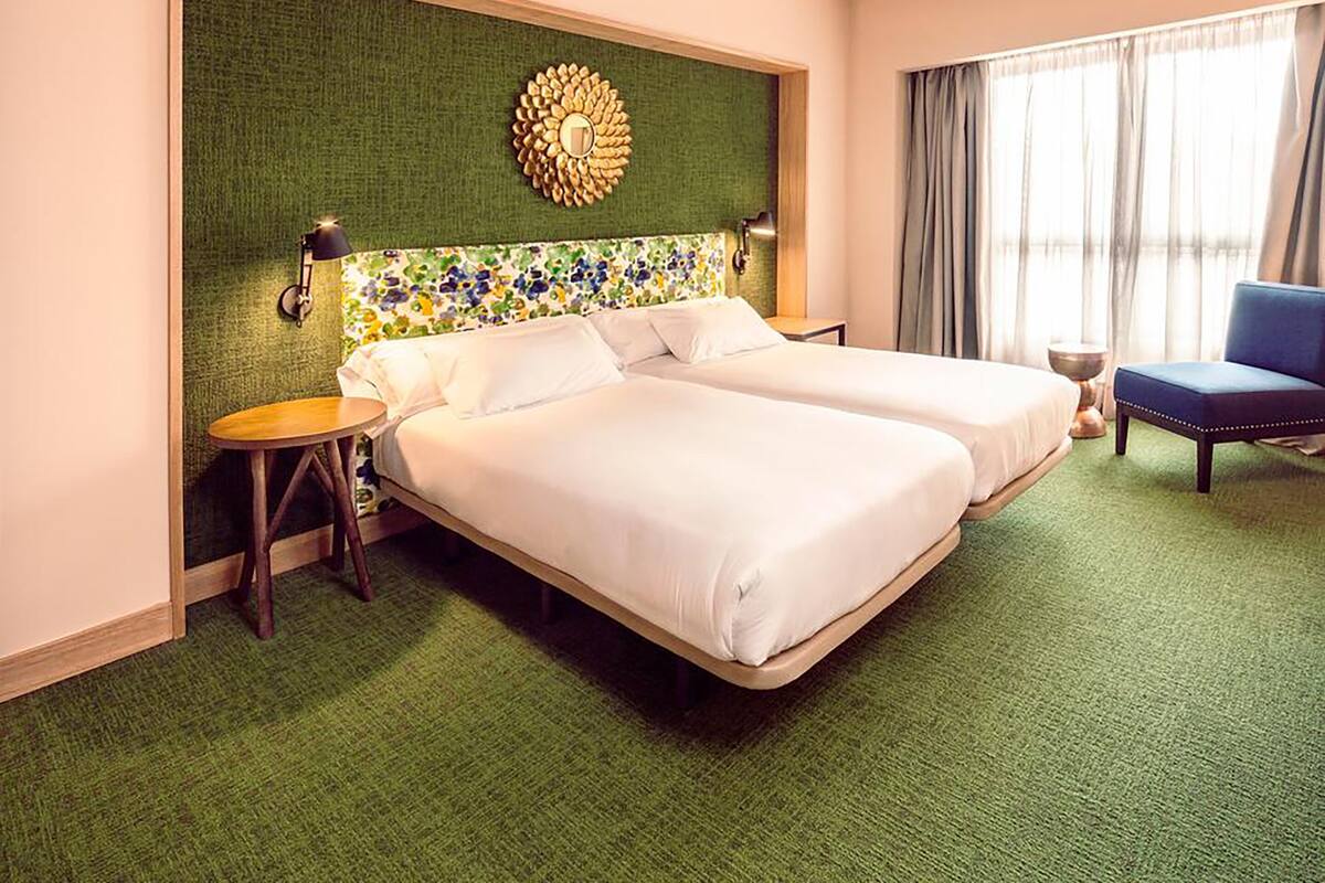 Habitación de hotel con dos camas, pared verde con textura, adorno de pared, una mesita redonda y una silla azul.