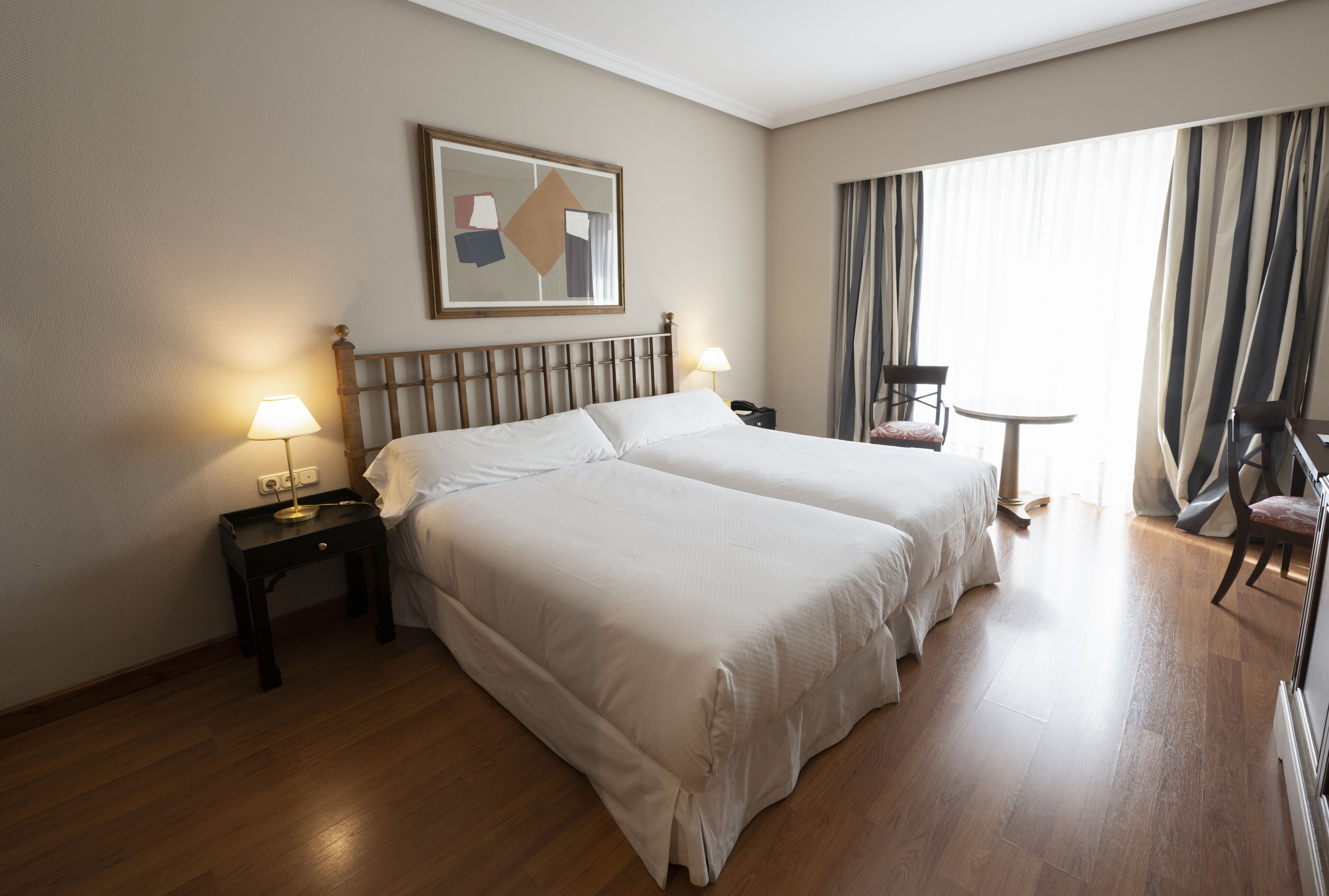 Een hotelkamer met twee bedden, een nachtkastje met lamp, een stoel, een klein tafeltje en een raam met gordijnen.