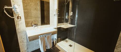 A modern bathroom with a large mirror, two sinks, a shower, and a hairdryer on the wall.