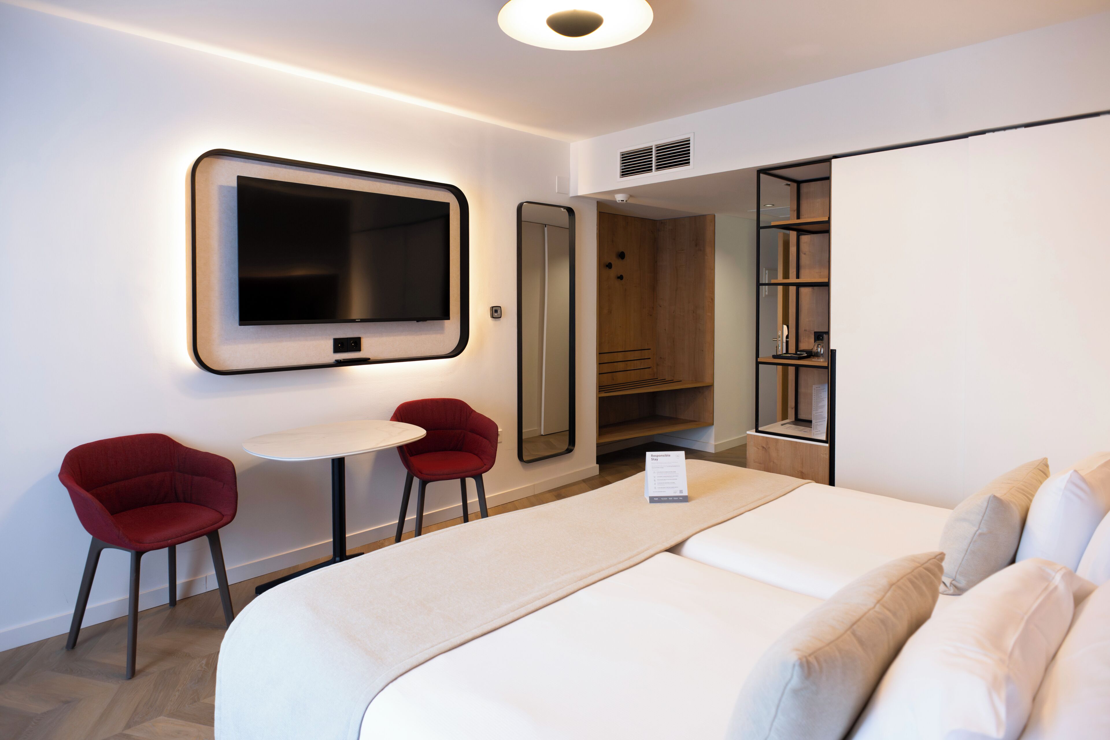 Une chambre d’hôtel avec un lit, deux chaises rouges, une petite table, un téléviseur, une armoire et un miroir.