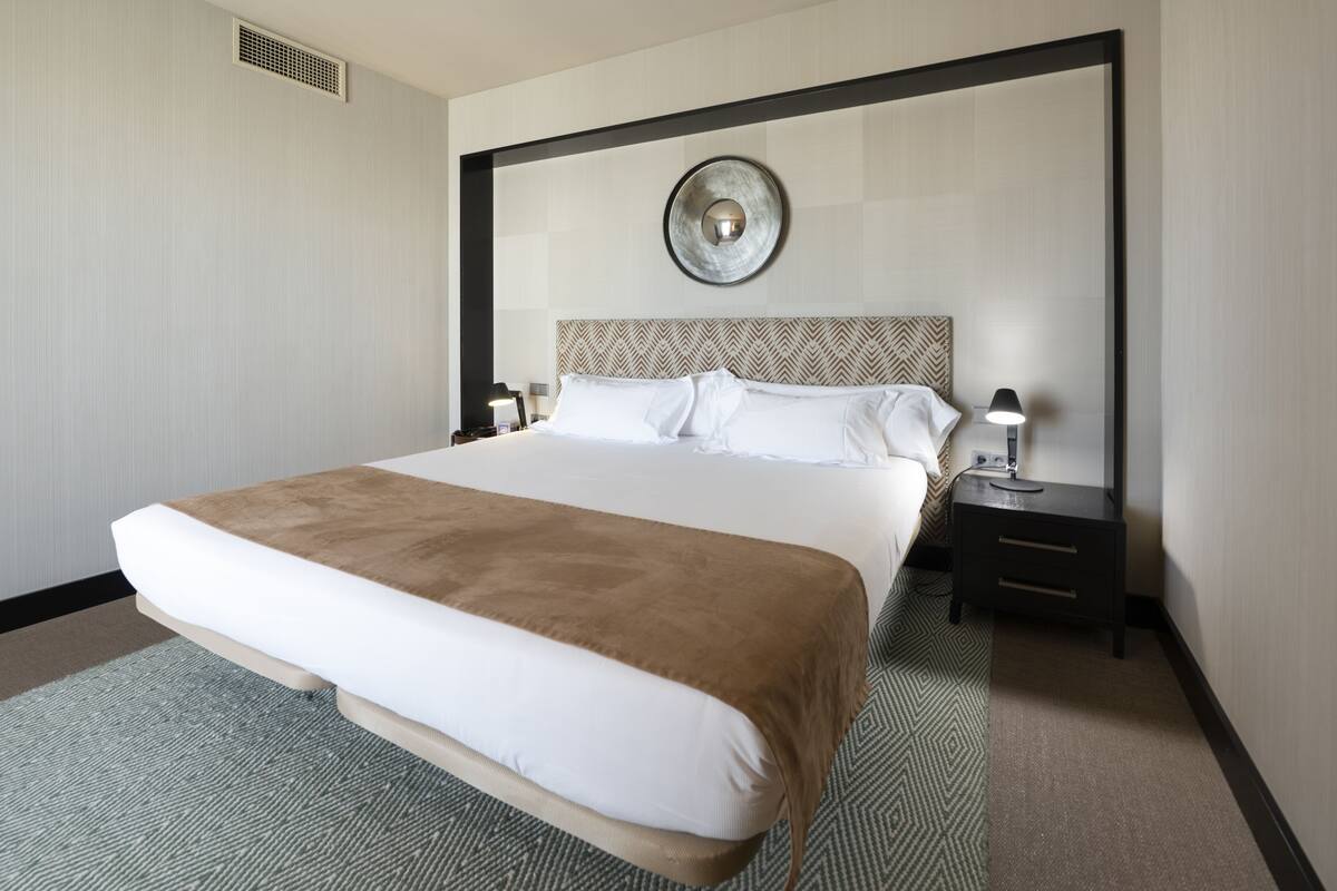 Habitación de hotel con una cama grande, una mesita de noche con lámpara y una lámpara de pared.