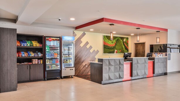 Zona de recepción con mostrador, refrigerador Coca-Cola, estantes con snacks y una pared decorada con un diseño geométrico.