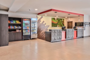 Zona de recepción con mostrador, refrigerador Coca-Cola, estantes con snacks y una pared decorada con un diseño geométrico.