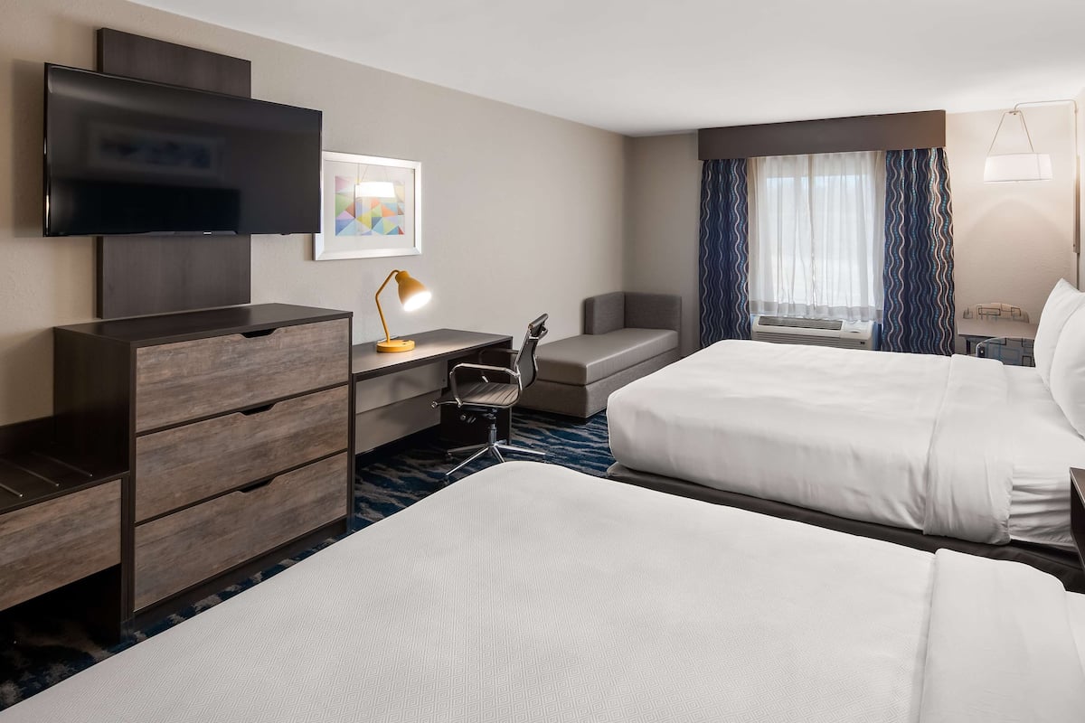 A hotel room with two beds, a desk with a chair, a TV, a lamp, and a window with curtains.
