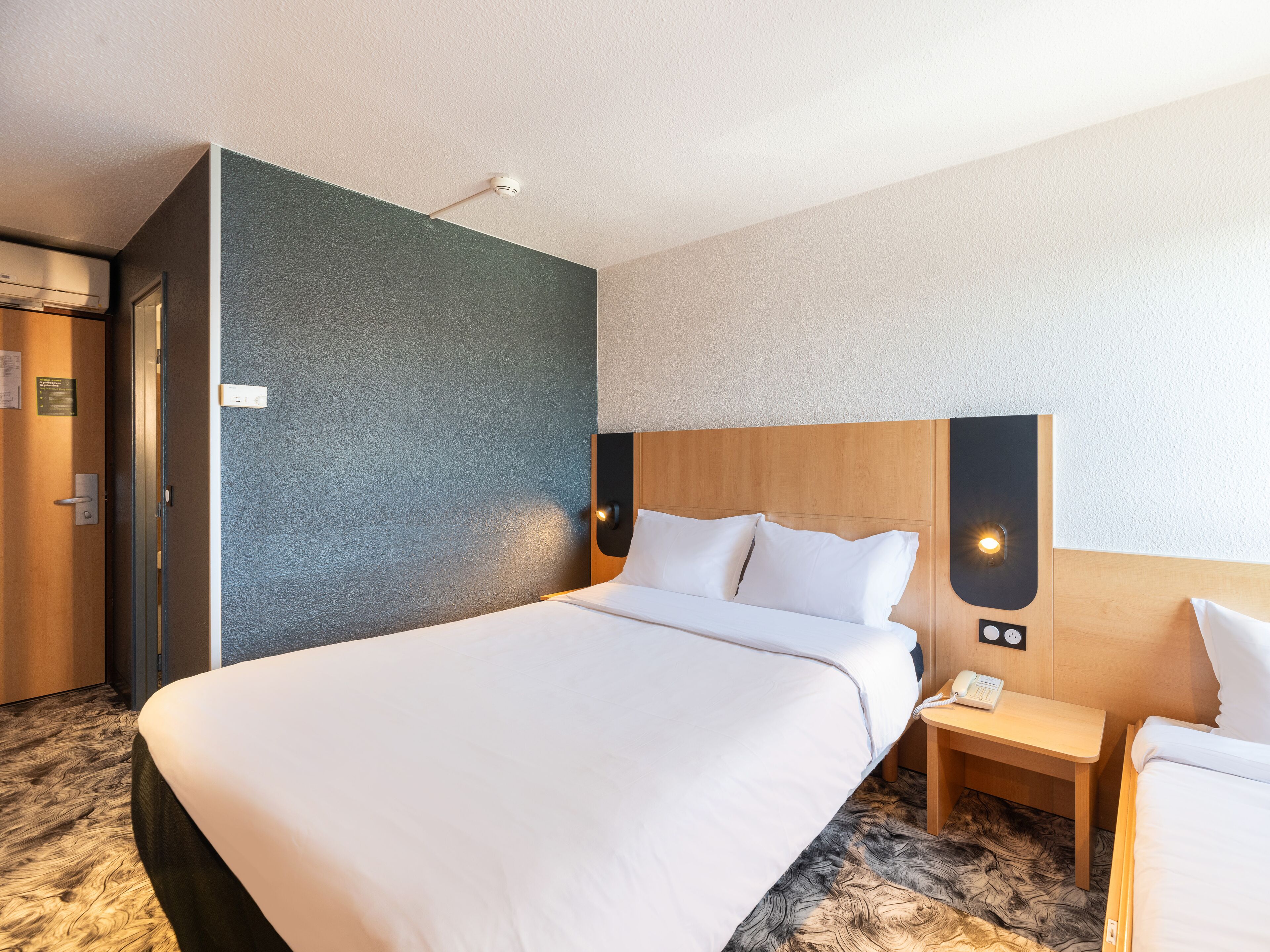 A hotel room with a large bed, a wooden nightstand with a phone, and a wall-mounted light fixture.