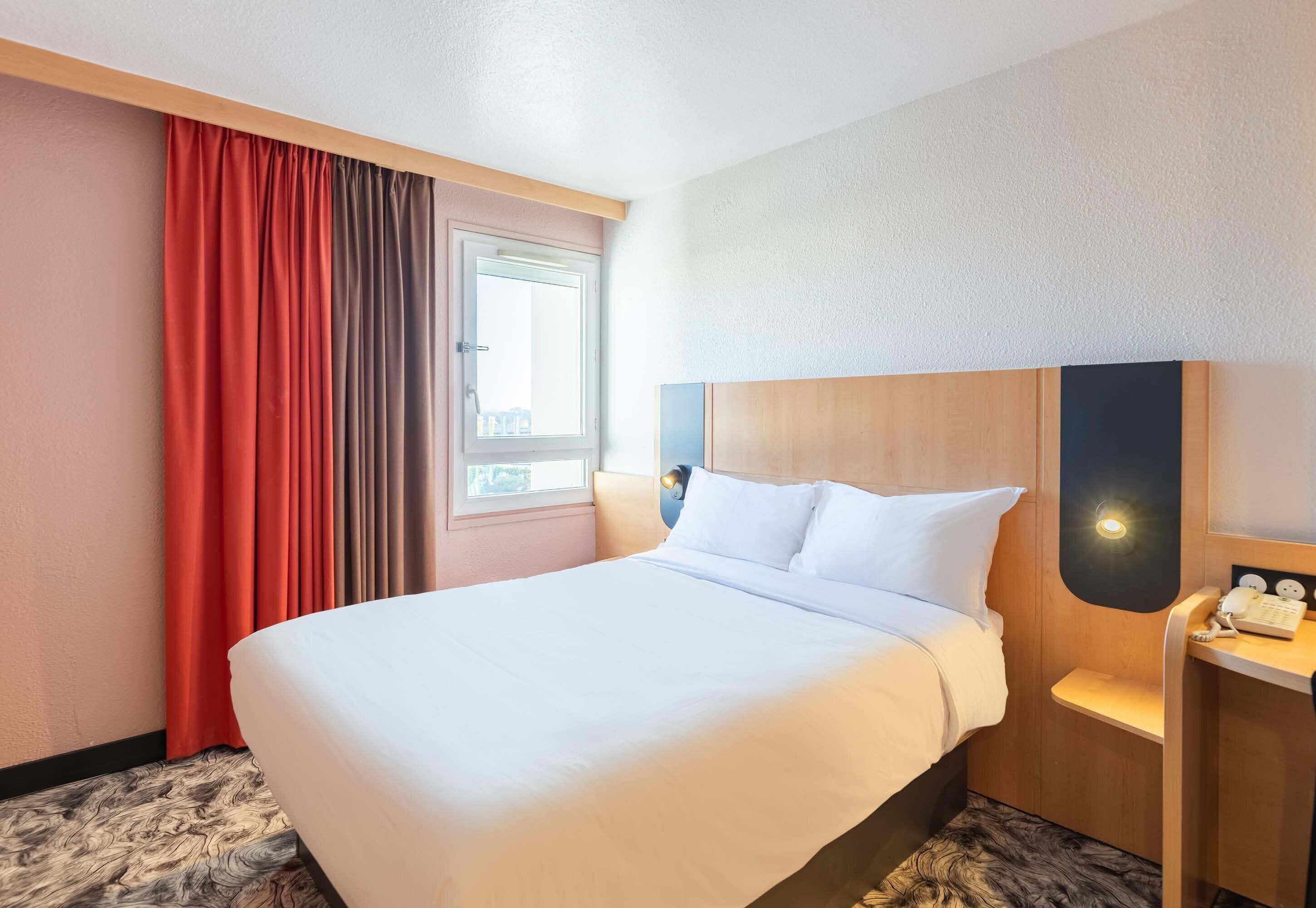Habitación de hotel con una cama grande, una mesita de noche, una lámpara, una ventana con cortinas y vistas al exterior.