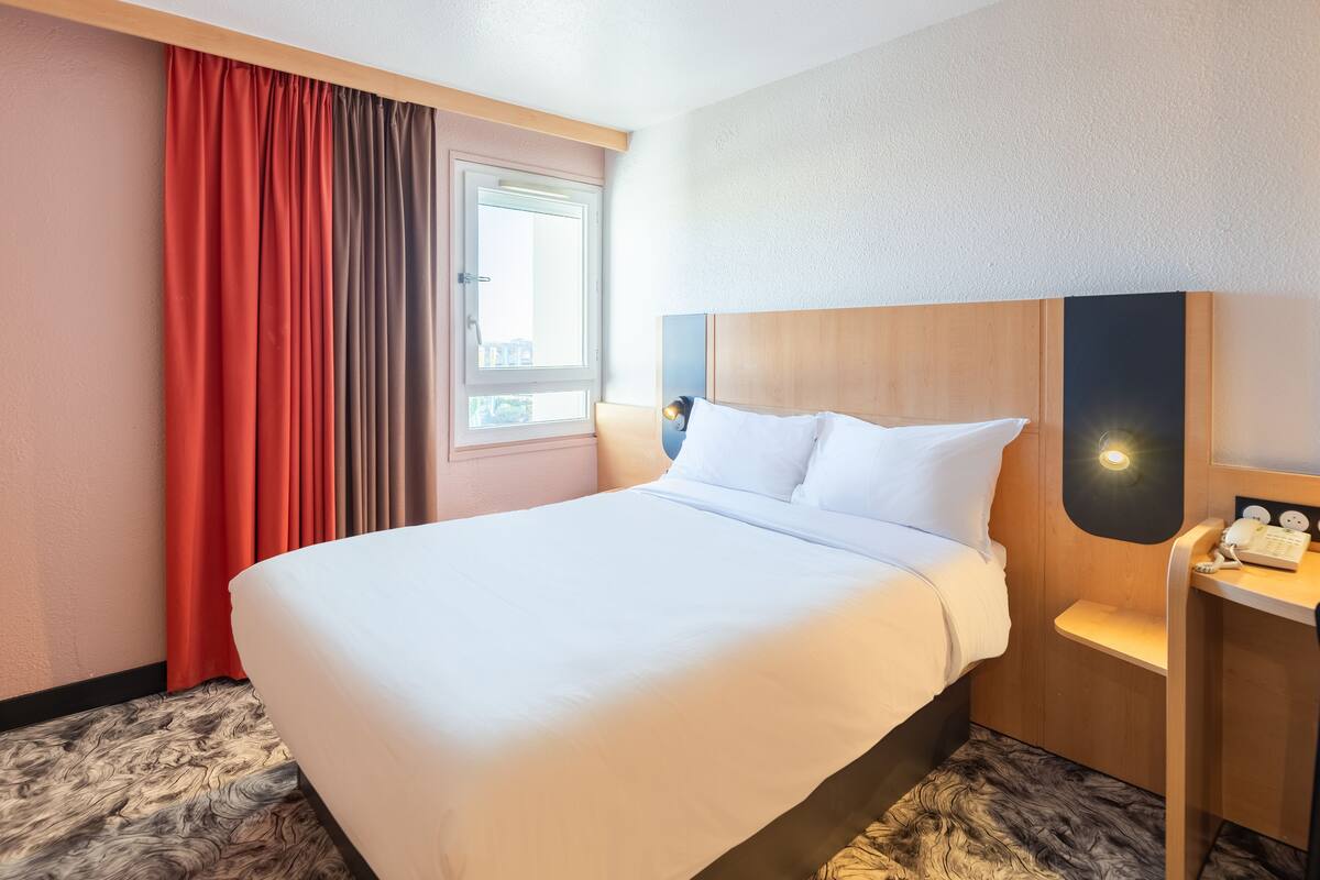 Una habitació d'hotel amb un llit gran, un capçal de fusta, una taula de nit amb una làmpada i una finestra amb cortines.