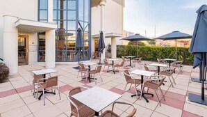 Een terras buiten met tafels, stoelen, parasols en een gebouwde ingang.