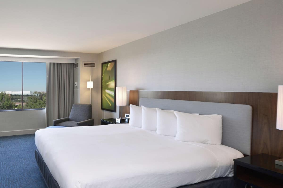 A hotel room with a large bed, a chair, a desk, and a window with a view of buildings.