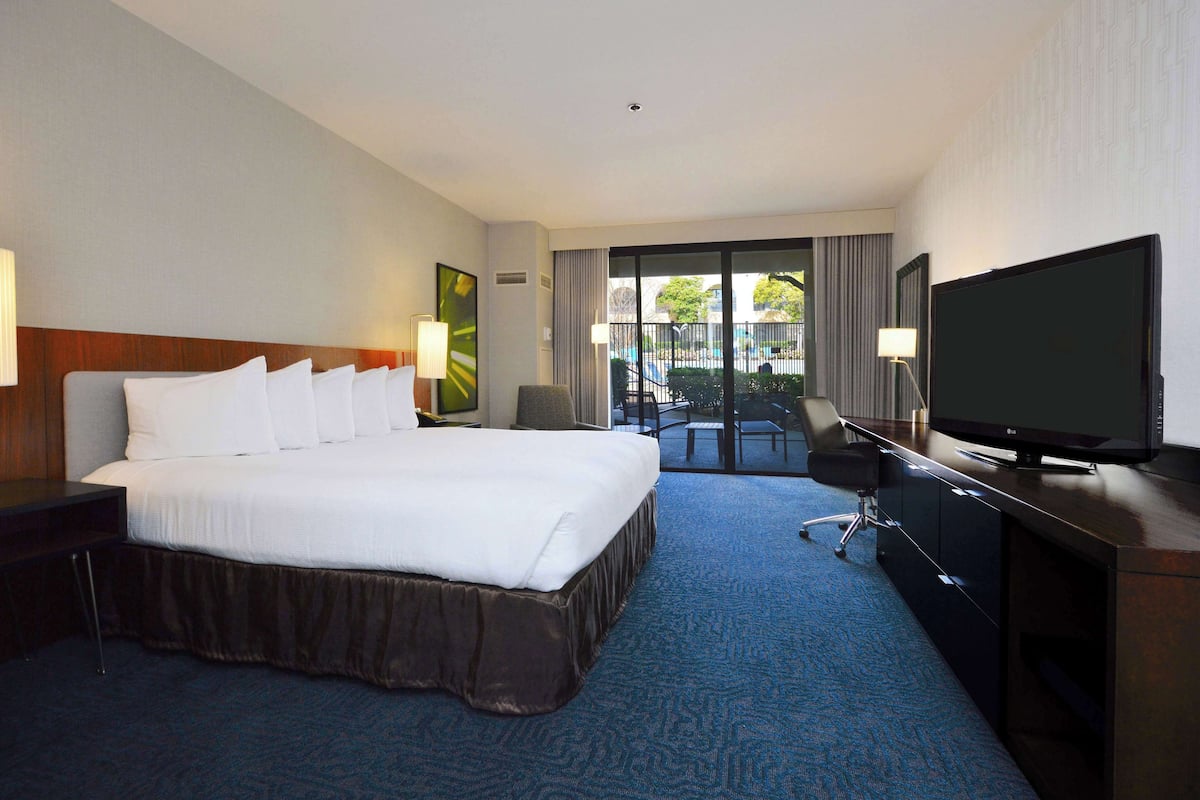 A hotel room with a large bed, a desk with a TV, and a view of a swimming pool.