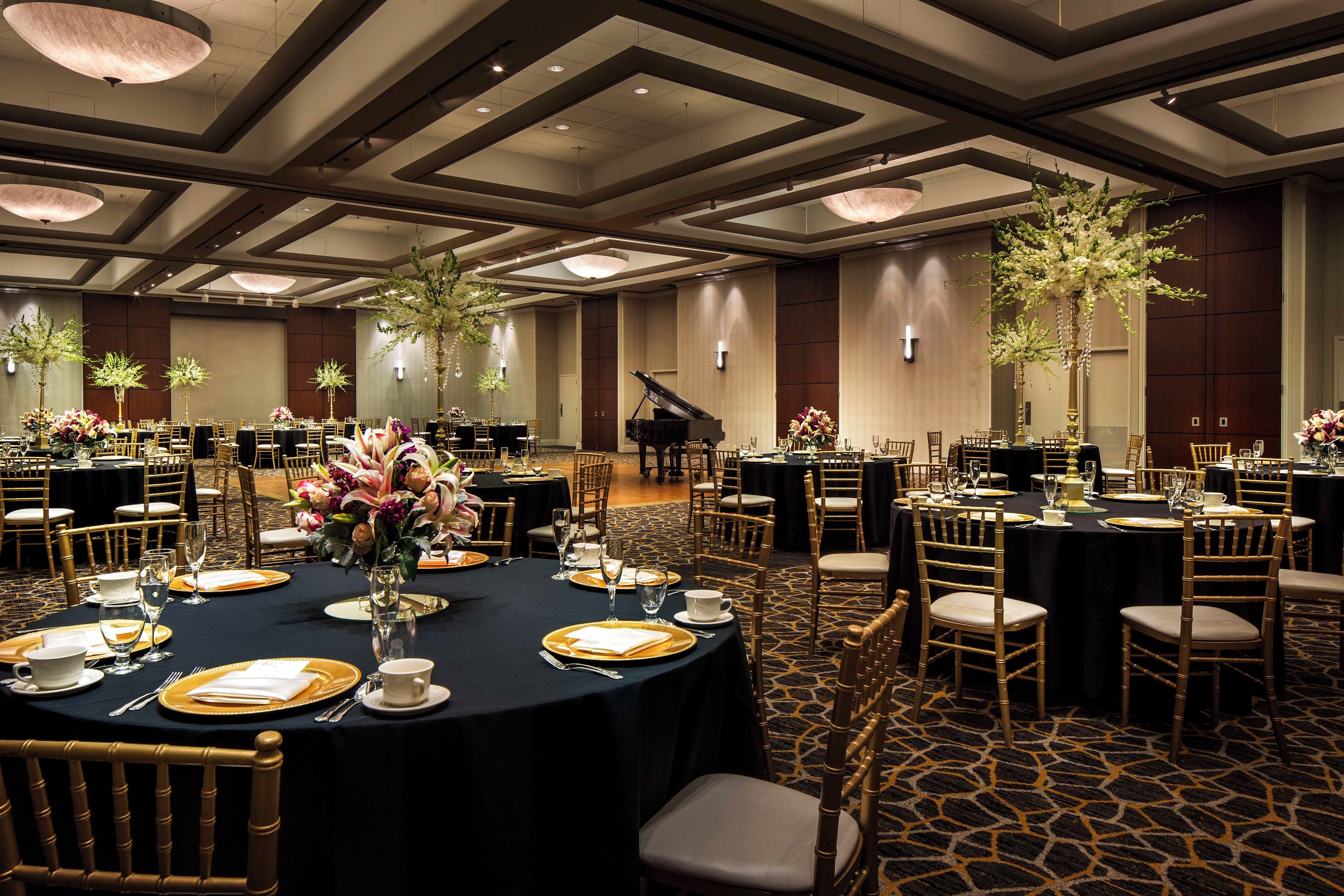 Een feestzaal met ronde tafels, gouden stoelen en bloemdecoraties.