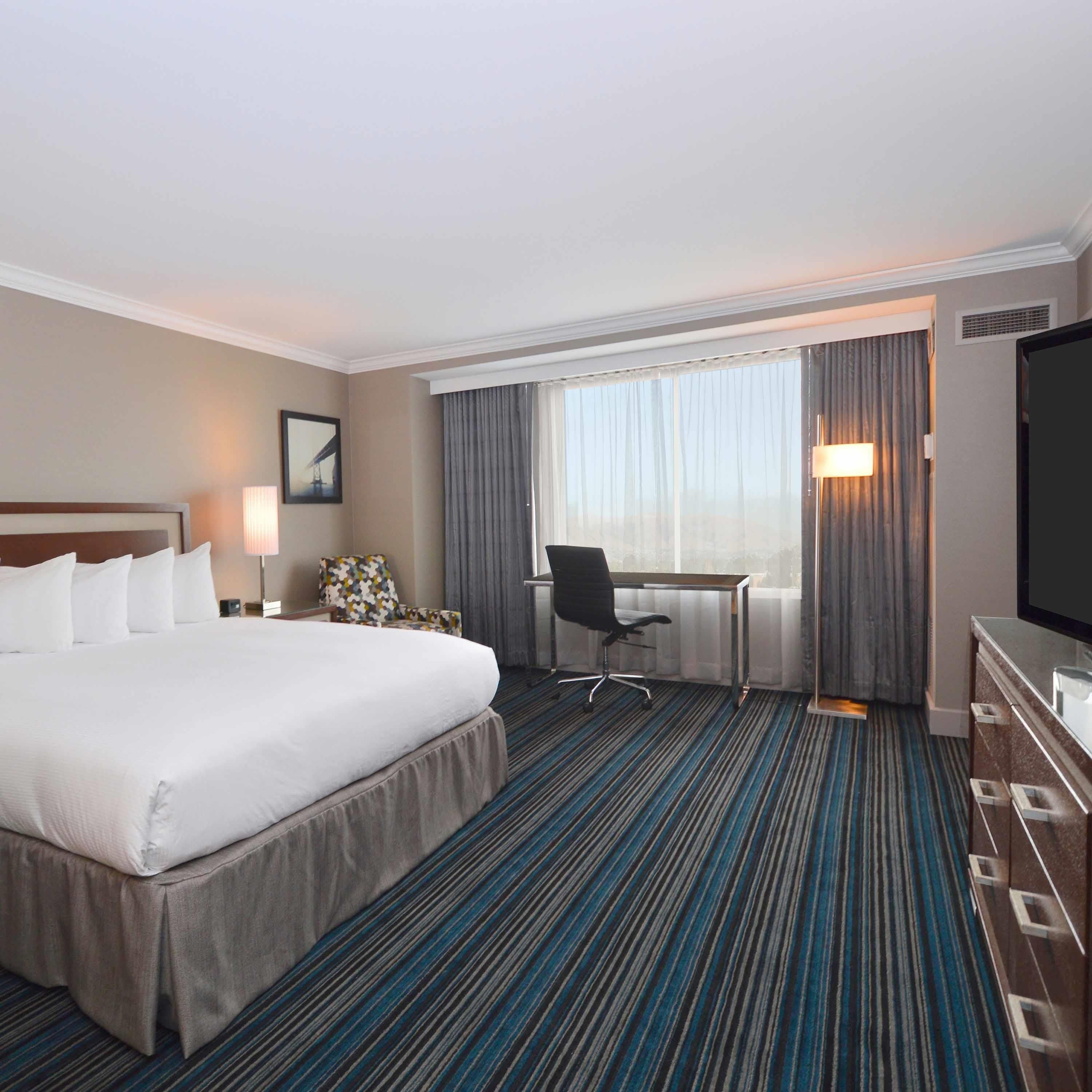 Ein Hotelzimmer mit einem großen Bett, einem Schreibtisch, einem Stuhl, einem Fernseher und einem Fenster mit Vorhängen.