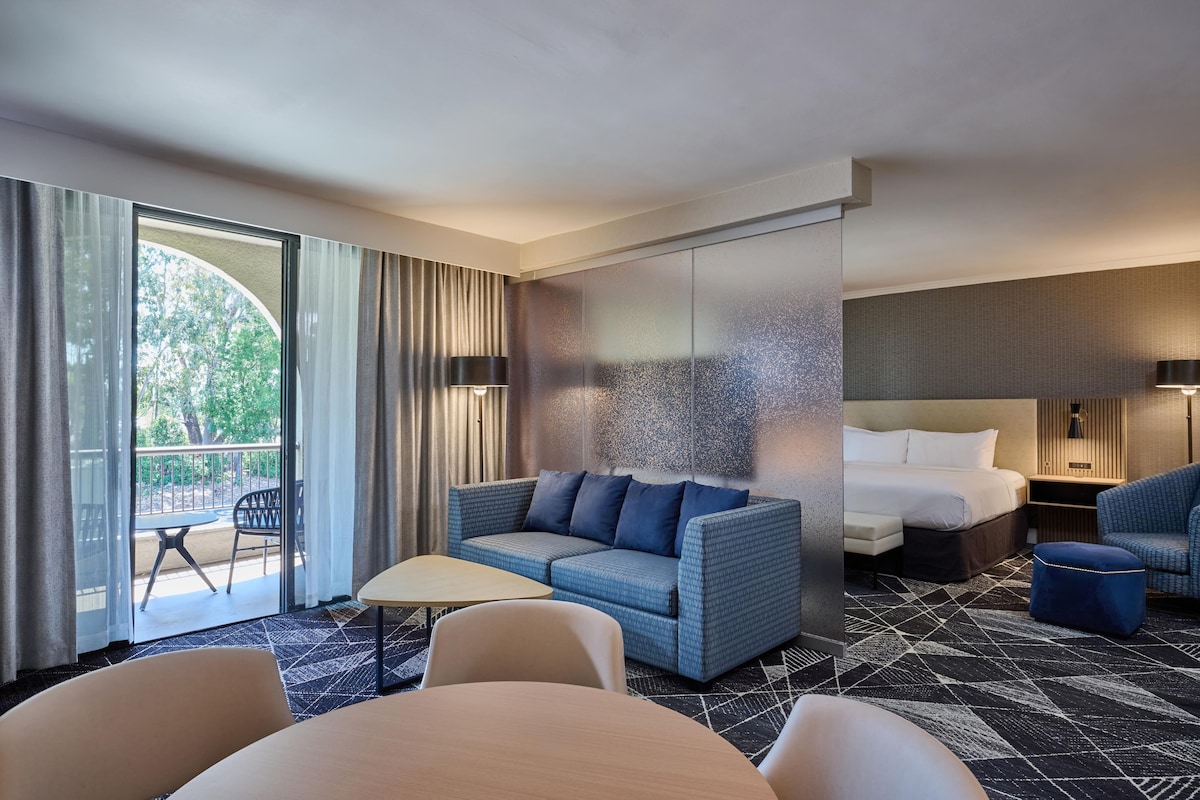 A hotel room with a sofa, a bed, a table, and a chair. There is a balcony with a view of trees.